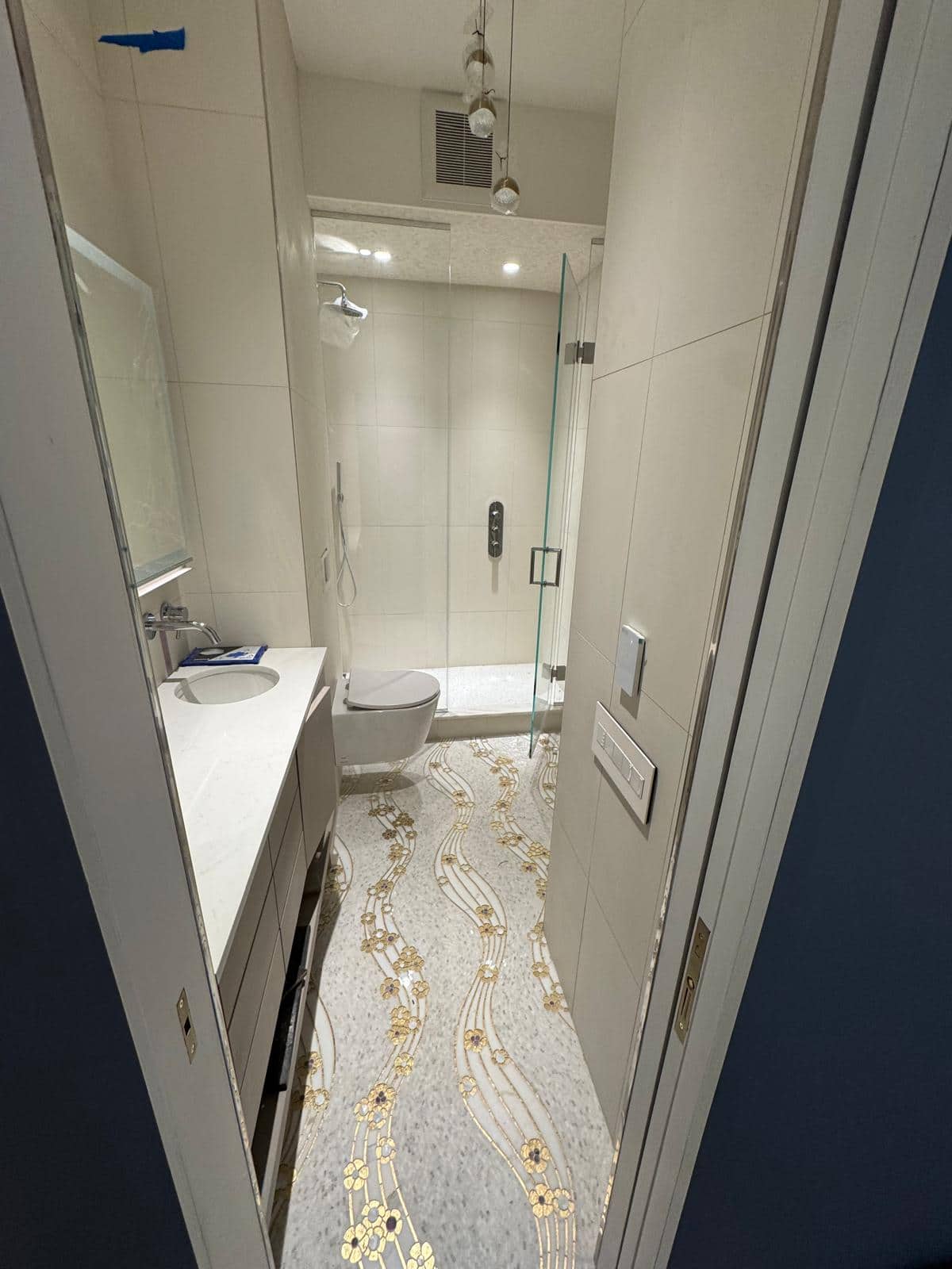 This narrow bathroom brings a white vanity, toilet, glass shower, pale wall tile, and patterned floor into one sight line.