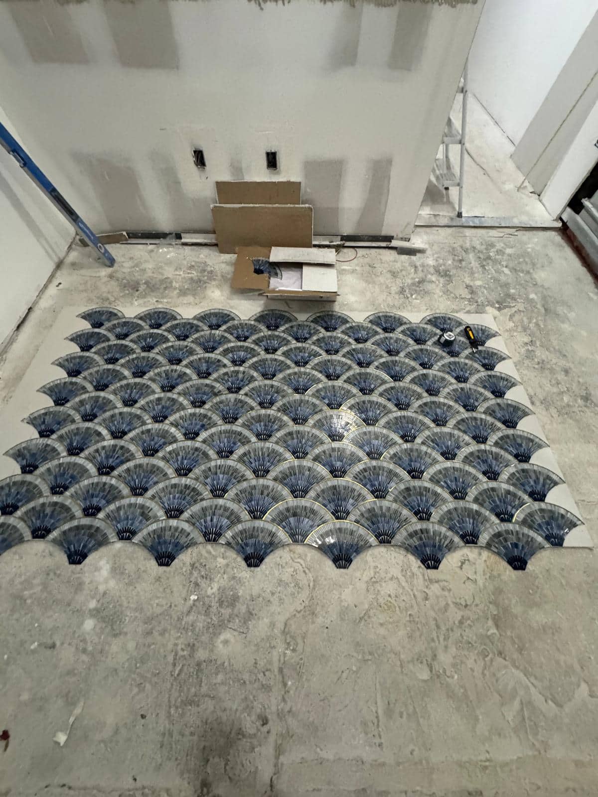 A section of blue scallop tile is laid out against an unfinished floor.